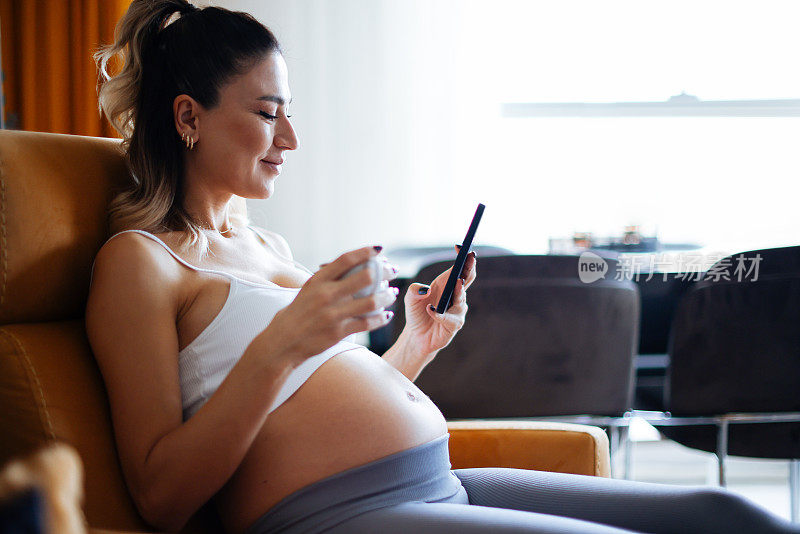 美丽的孕妇喝咖啡/茶，在家里玩手机。